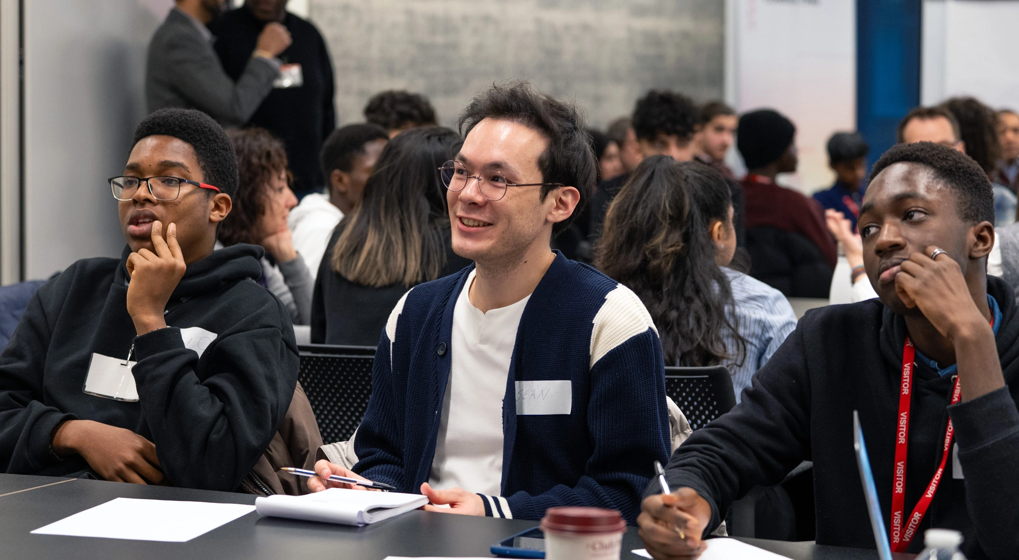 Three students sit at a desk listening to a speaker